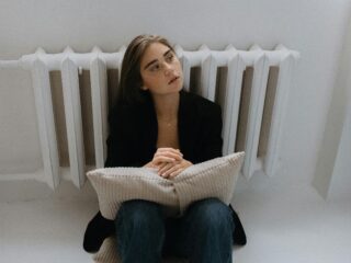 Osoba sjedi na podu uz radijator i drži jastuk, u tihoj pauzi bez distrakcija.