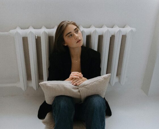 Osoba sjedi na podu uz radijator i drži jastuk, u tihoj pauzi bez distrakcija.